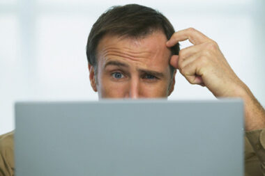 Man looking confused scratching his head behind a laptop