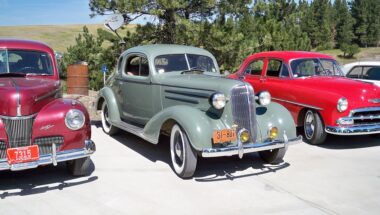 Some Classic Cars lined up in a car park with trees in the background