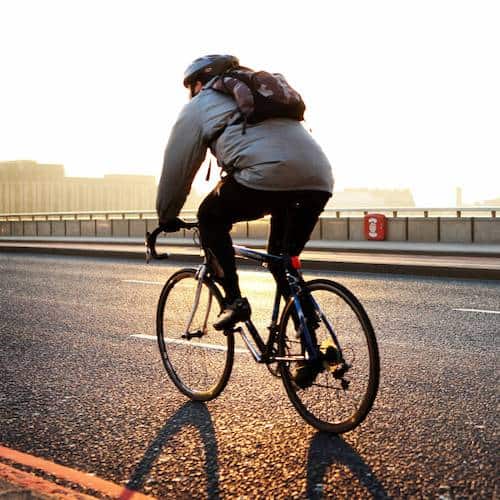Back view of a man wearing a backpack riding a bicycle alone a road with the sun setting in the background
