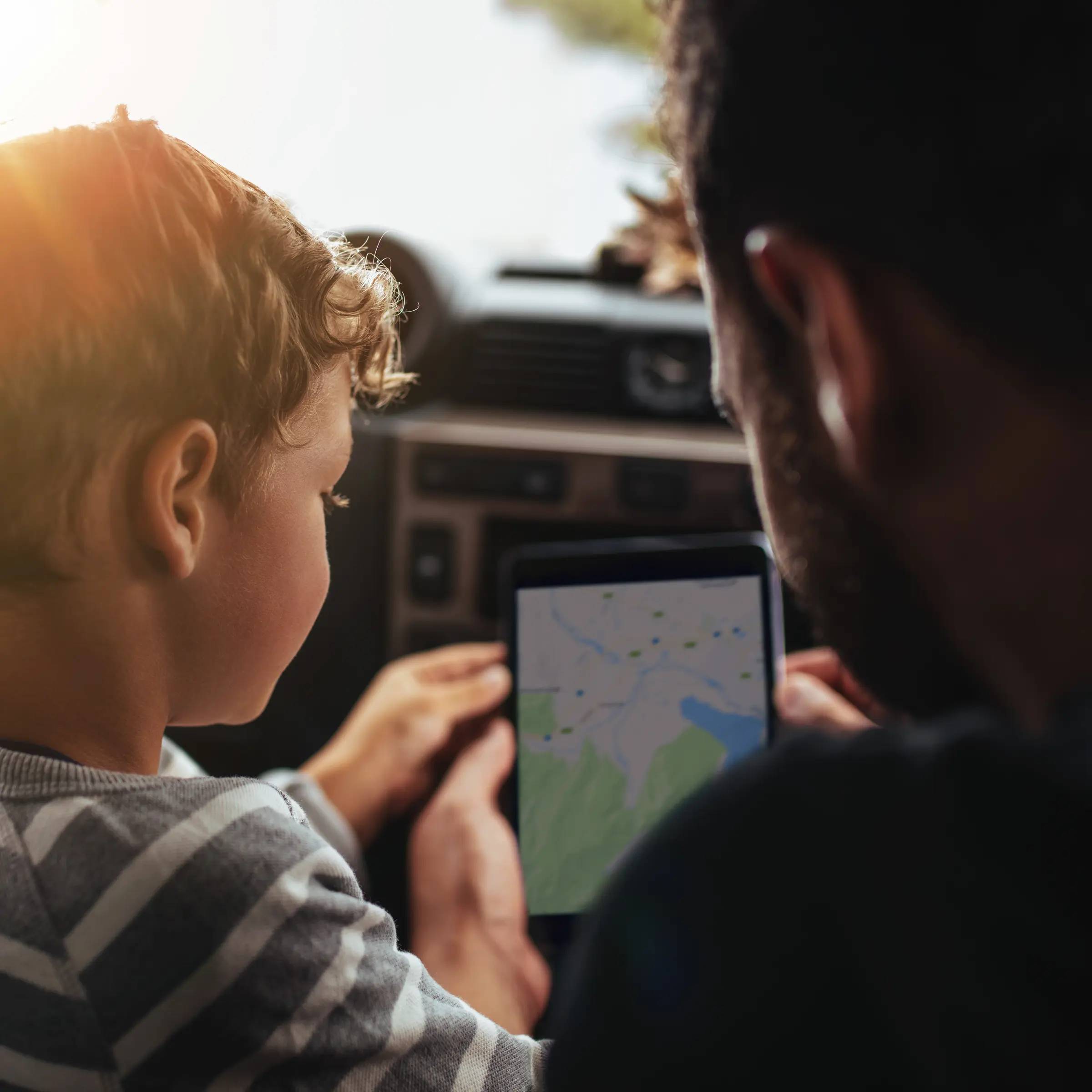 A father and young son are in the front of a parked car, looking at a map on a tablet. 