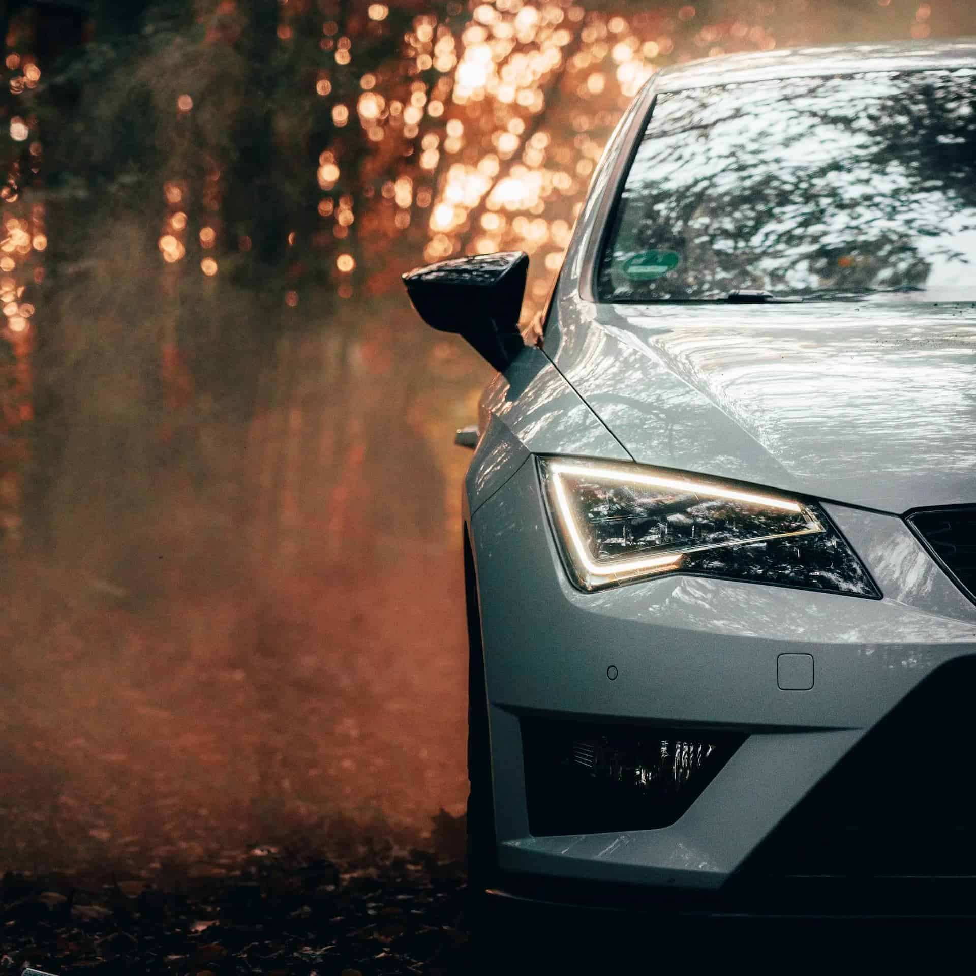 Front left of a white car with headlight illuminated with autumn trees and fog