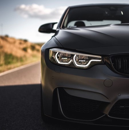 The front end of a black matte sports car on a countryside road. 