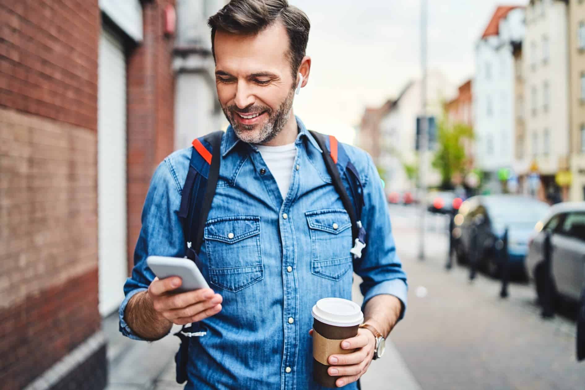 A man in a denim jacket walking down a street. He is holding a takeaway coffee in one hand while looking at the mobile in the other hand.