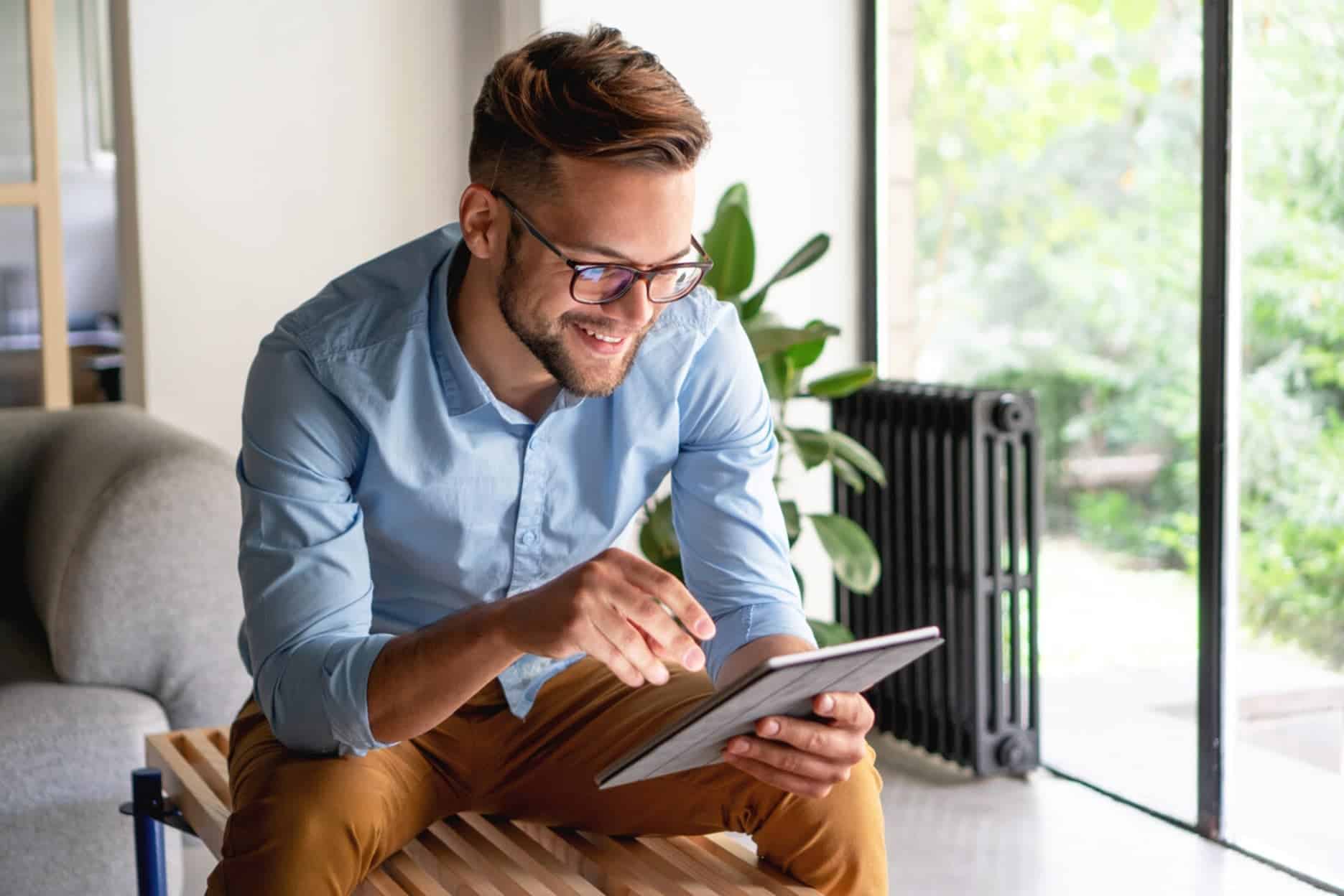 A smartly dressed man sits on an indoor wooden bench while using a tablet.