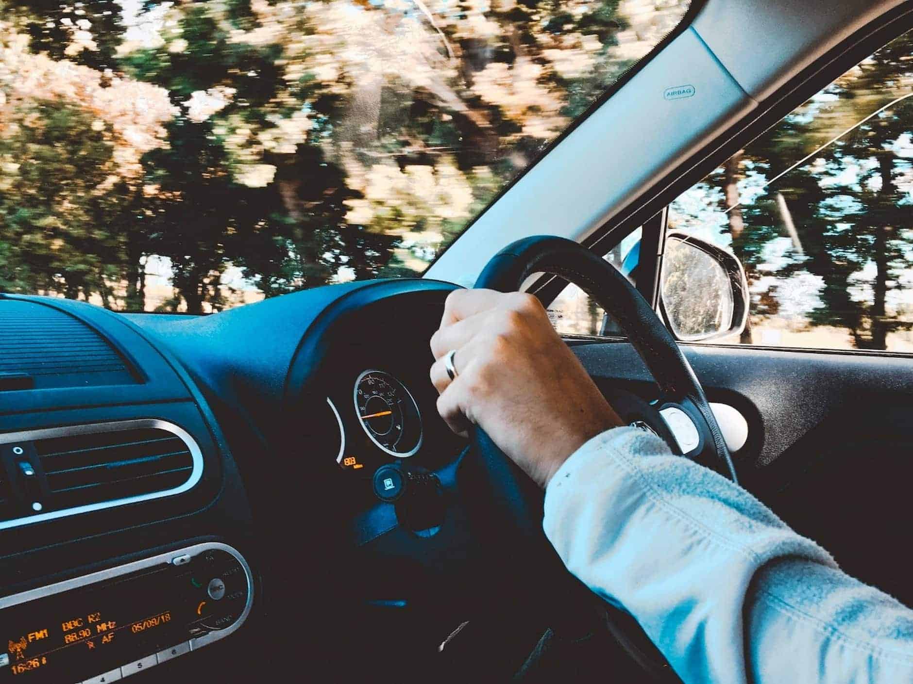 A man driving a car past a wooded landscape; the weather looks sunny 