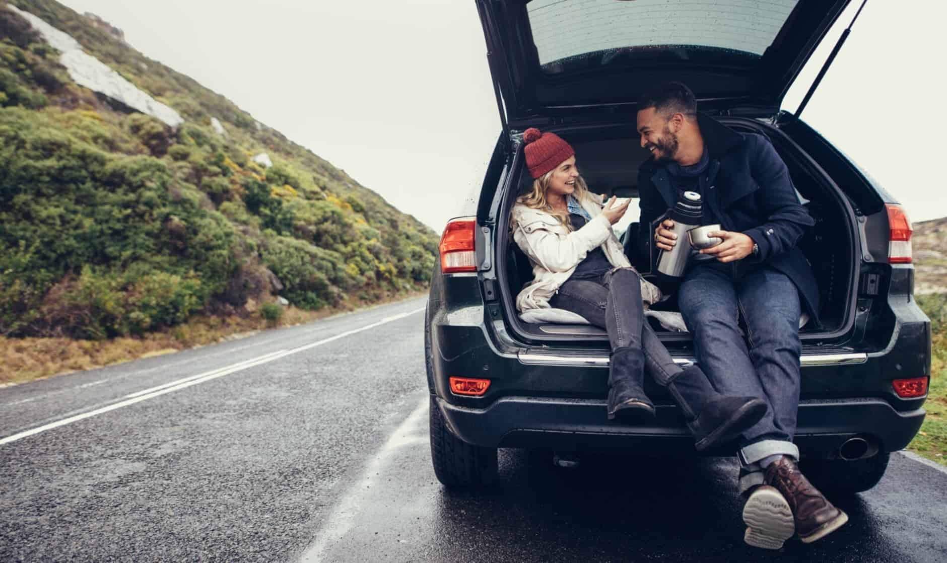 A man and a woman laughing in the boot of their car, enjoying a hot drink whilst parked on the side of a country road.