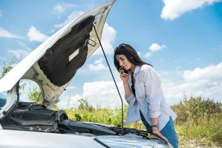 Young woman looks under the engine hood as broken cars motor